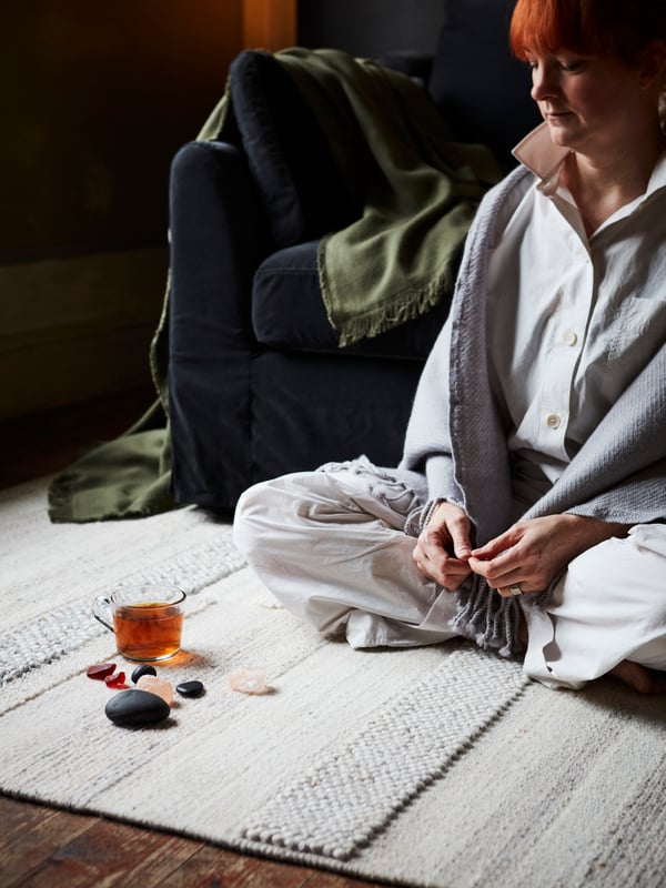 Eine Person sitzt auf einem hellen Teppich in einem Wohnzimmer. Vor ihr liegen Kristalle, Steine und ein Becher aus Glas.