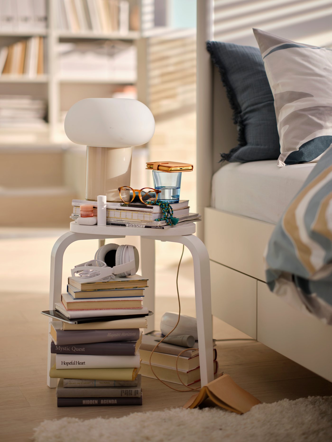 A white KYRRE stool with a DEJSA table lamp, a glass of water, a mobile phone and other things on it is placed next to a bed.