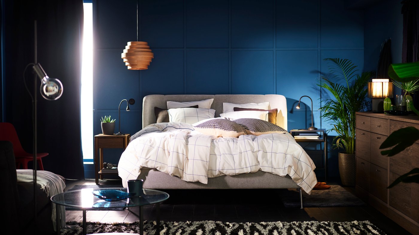 A TUFJORD upholstered bed in a blue bedroom covered with BREDVECKLARE bed linen. A DYKARKLOCKA pendant lampshade hangs above.
