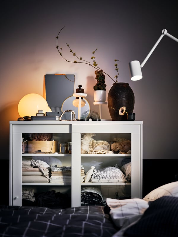 Près d’un lit se trouve une vitrine SYVDE blanche remplie de rangement, avec des décorations et une lampe de table FADO blanche allumée sur le dessus.