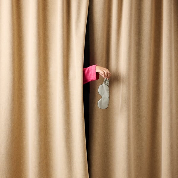 A person’s hand is sticking out from behind pulled ROSENMANDEL blackout curtains, about to drop a sleep mask on the floor.