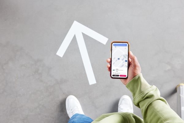 Osoba u zelenoj majici drži mobilni telefon s prikazom karte robne ku'e IKEA. Na sivom podu vidljiva je bela strelica koja pokazuje smer kretanja.