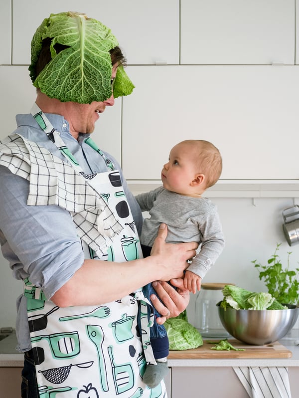 Homme debout dans une cuisine portant un tablier RINNIG, une feuille de chou sur la tête. Il porte un bébé qui a l’air surpris.