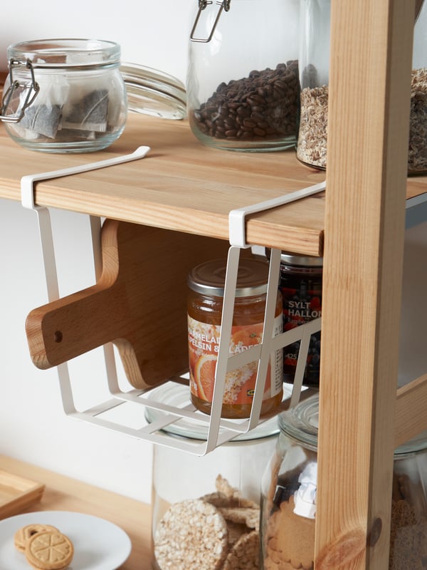 A PÅLYCKE clip-on basket attached to a wooden shelf, the basket holds a wooden chopping board and a jar, items on the shelf.
