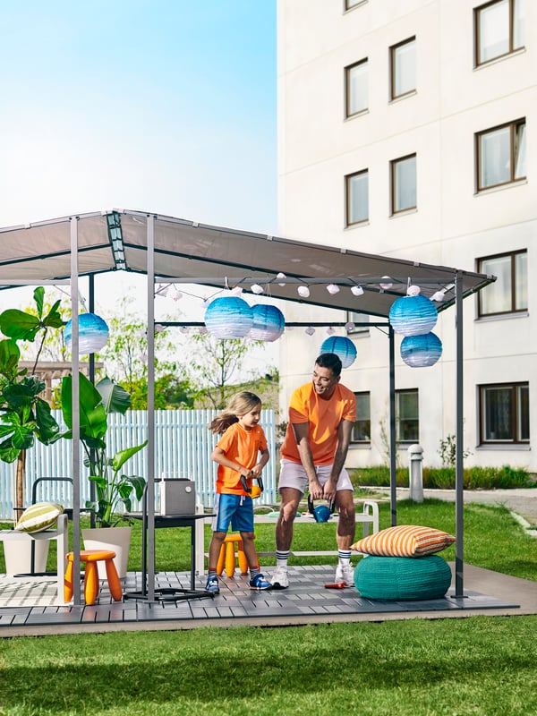 A summery backyard, a girl and father lifting weights in a GUNNÖN gazebo with RUNNEN outdoor floor decking, lamps and plants.