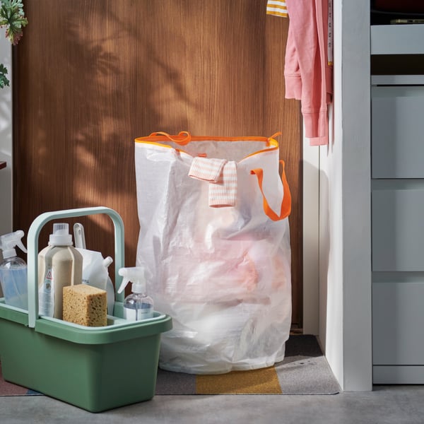 A hallway with a green PEPPRIG cleaning bucket and caddy beside a large white PÄRKLA laundry bag filled with clothes.