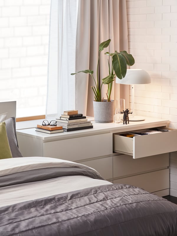 A white MALM chest of drawers with a comfy bed next to it and a plant, some books and a white NYMÅNE table lamp on top.