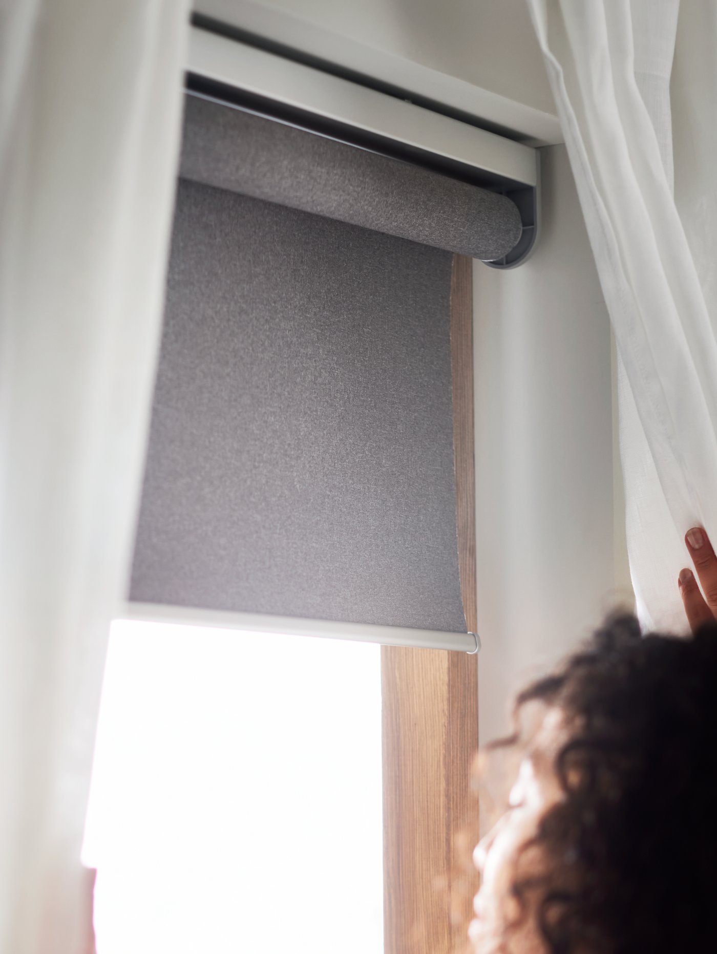 Una mujer junto a una ventana con cortinas blancas y una persiana enrollable gris FYRTUR que funciona con batería y es inalámbrico.