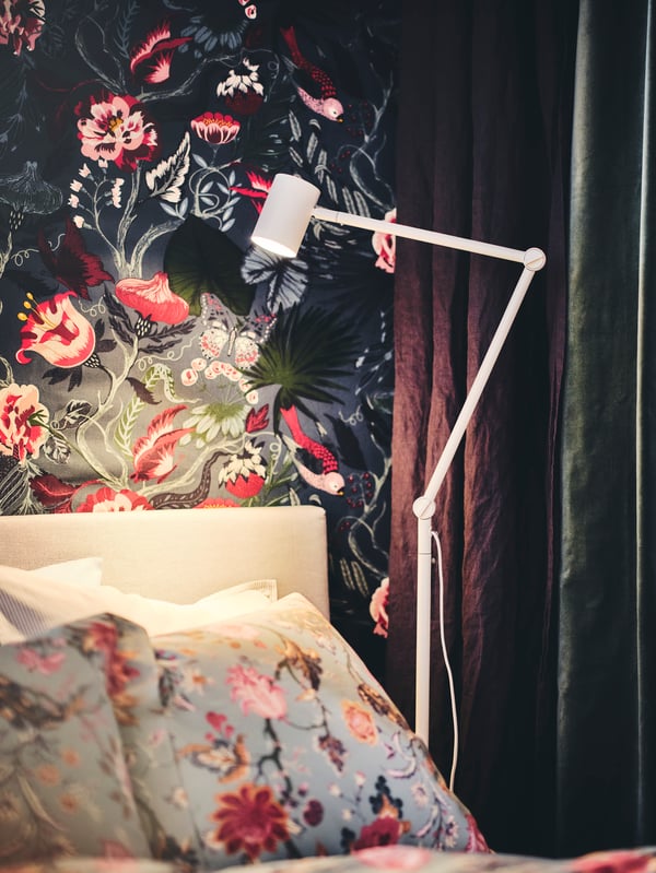 A white NYMÅNE floor/read lamp in a bedroom with a floral wall behind the bed, which is covered in NÄSSELKLOCKA bed linen.