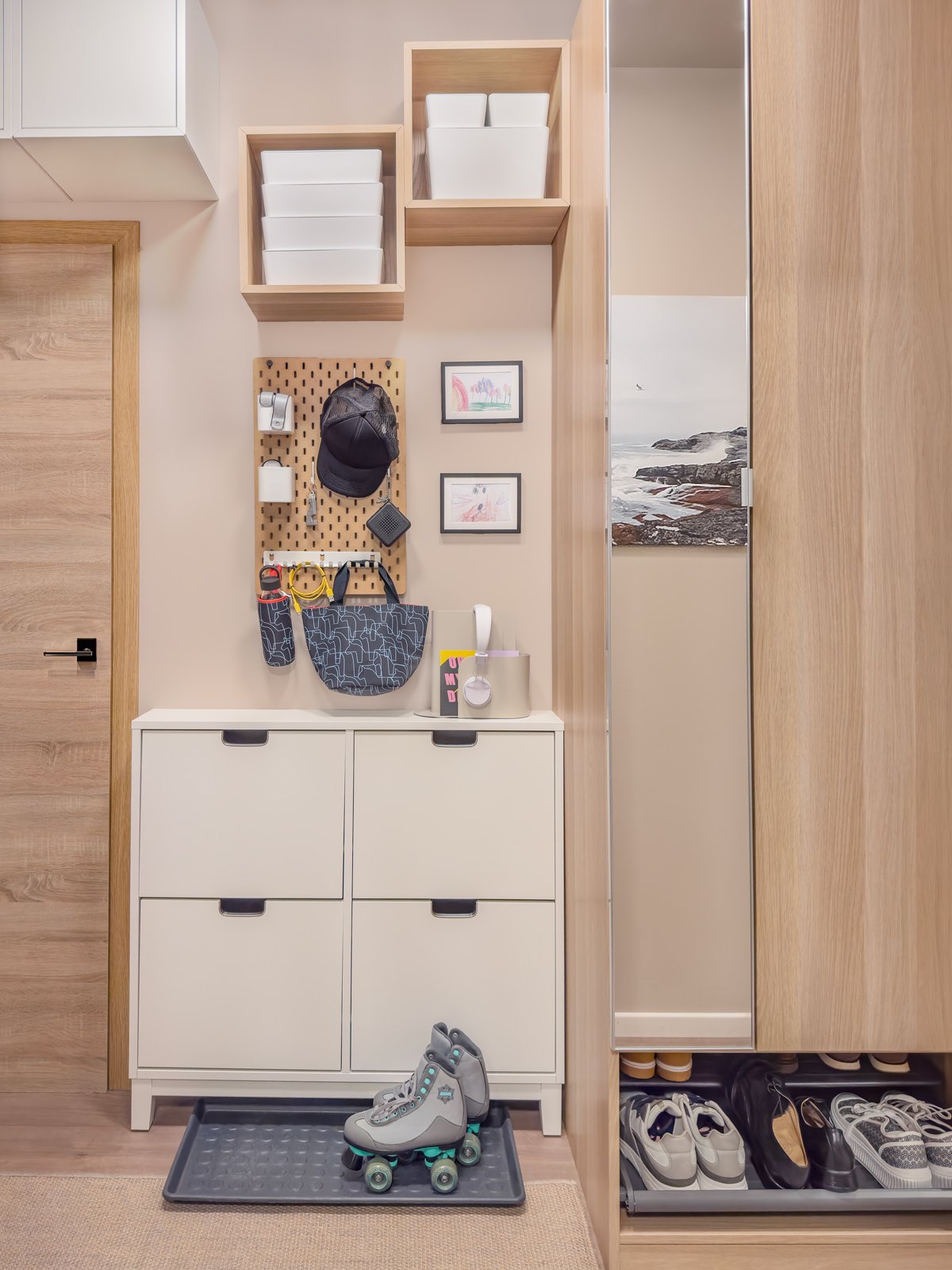 Entré med vit skoförvaring, hyllor med lådor, en perforerad väggpanel med krokar och små ramar, samt en spegelgarderob med skor och ett par rullskridskor på en matta.
