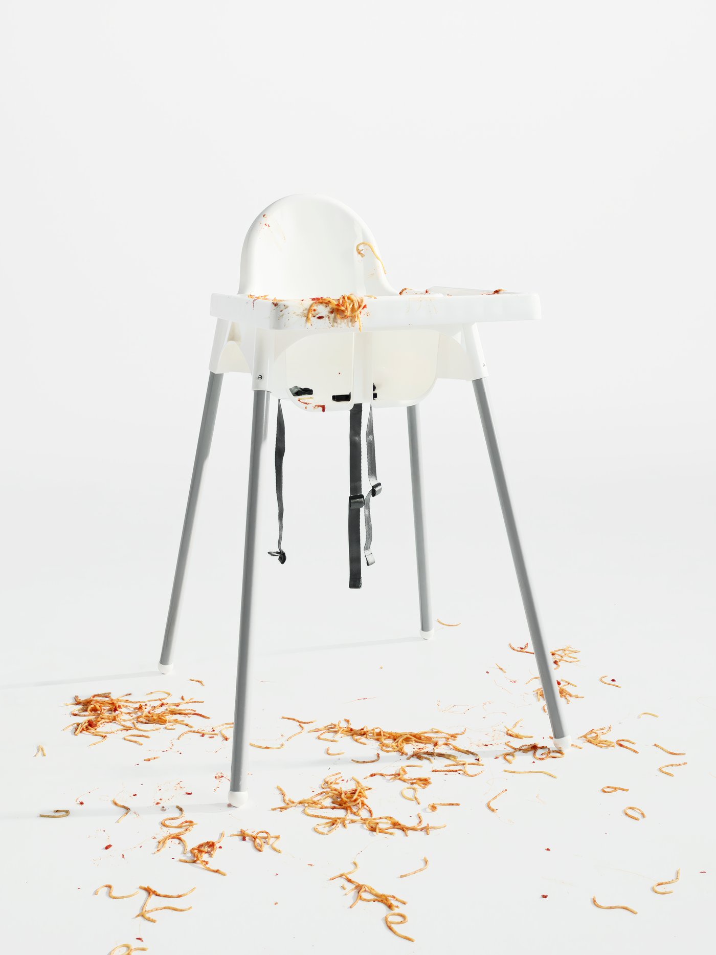 An empty highchair with tray in a white space. The remains of a messy spaghetti meal cover the highchair and floor.