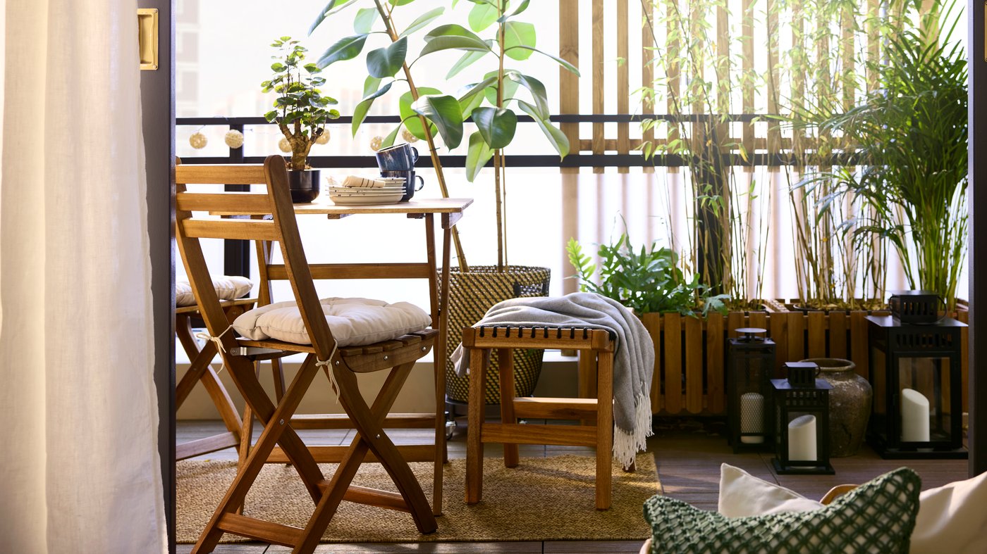 A light brown stained acacia ASKHOLMEN table and foldable chair face the view in a balcony with plants and natural materials.