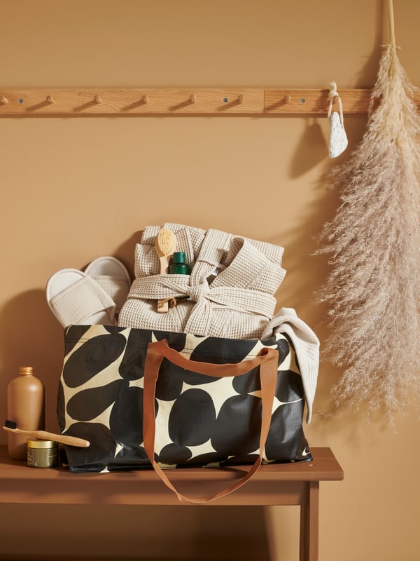 An off-white/black SÄCKKÄRRA carrier bag containing a bath robe, slippers and other items for guests sits on a bench.