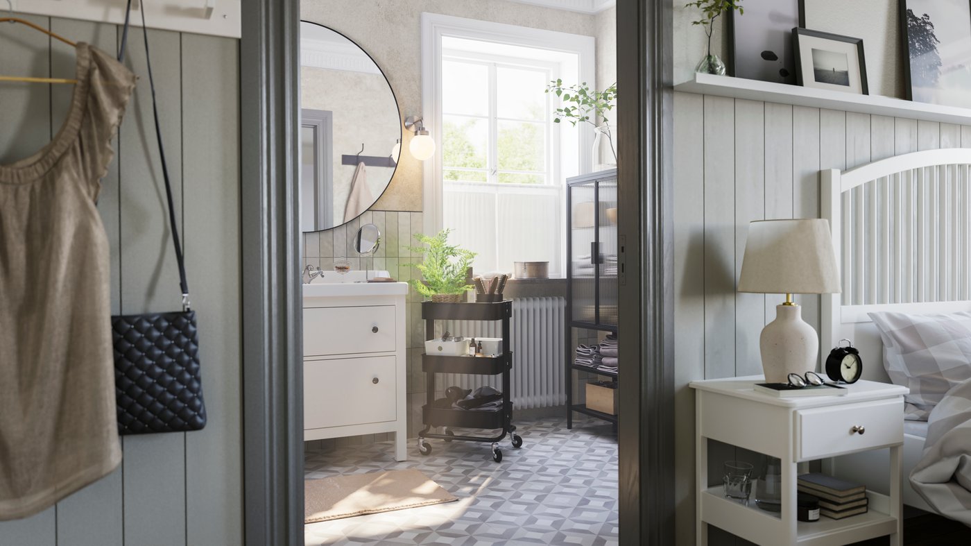 큰 창이 있는 회색 욕실에 HEMNES 헴네스 화이트 세면대와 RÅSKOG 로스코그 블랙 카트, MOSSJÖN 모셴 유리도어수납장이 있는 모습.