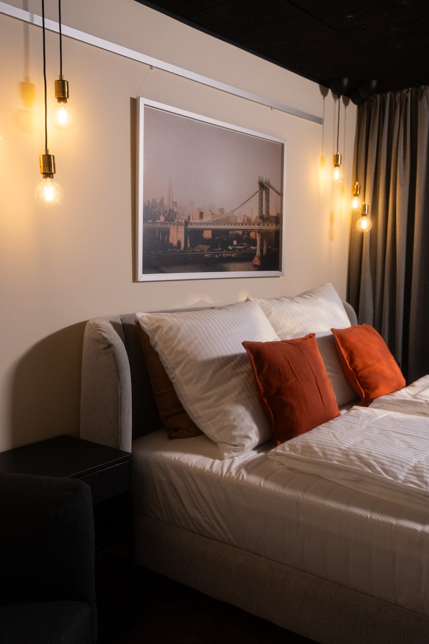 Bed with white sheets and pillows, decorated with rust-colored decorative pillows, warm lighting from hanging lamps and a painting above the bed.