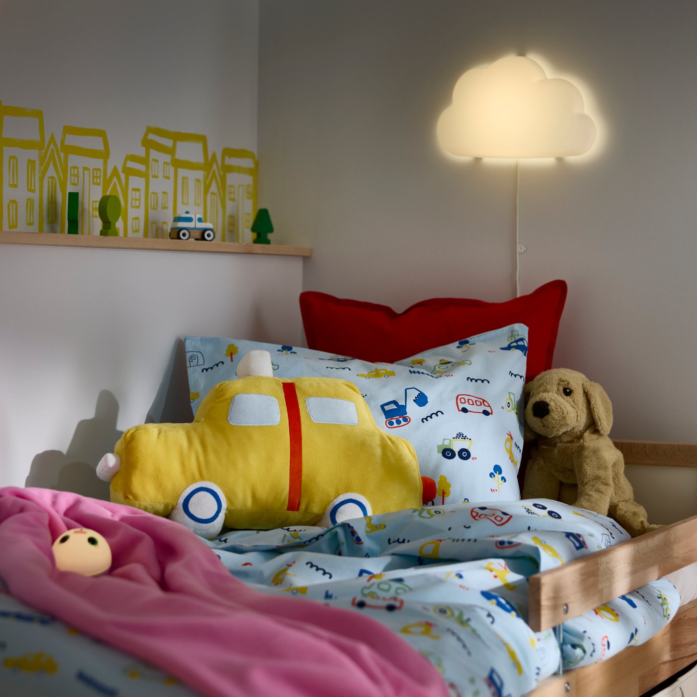 A children’s bed with the BARNDRÖM cushion shaped like a yellow car placed on patterned bedding with toys nearby.