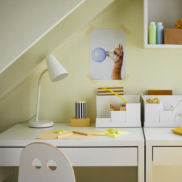 A FUBBLA work lamp and TJENA desk organisers filled with supplies are on top of two MICKE desks. Markers are on the desktop.