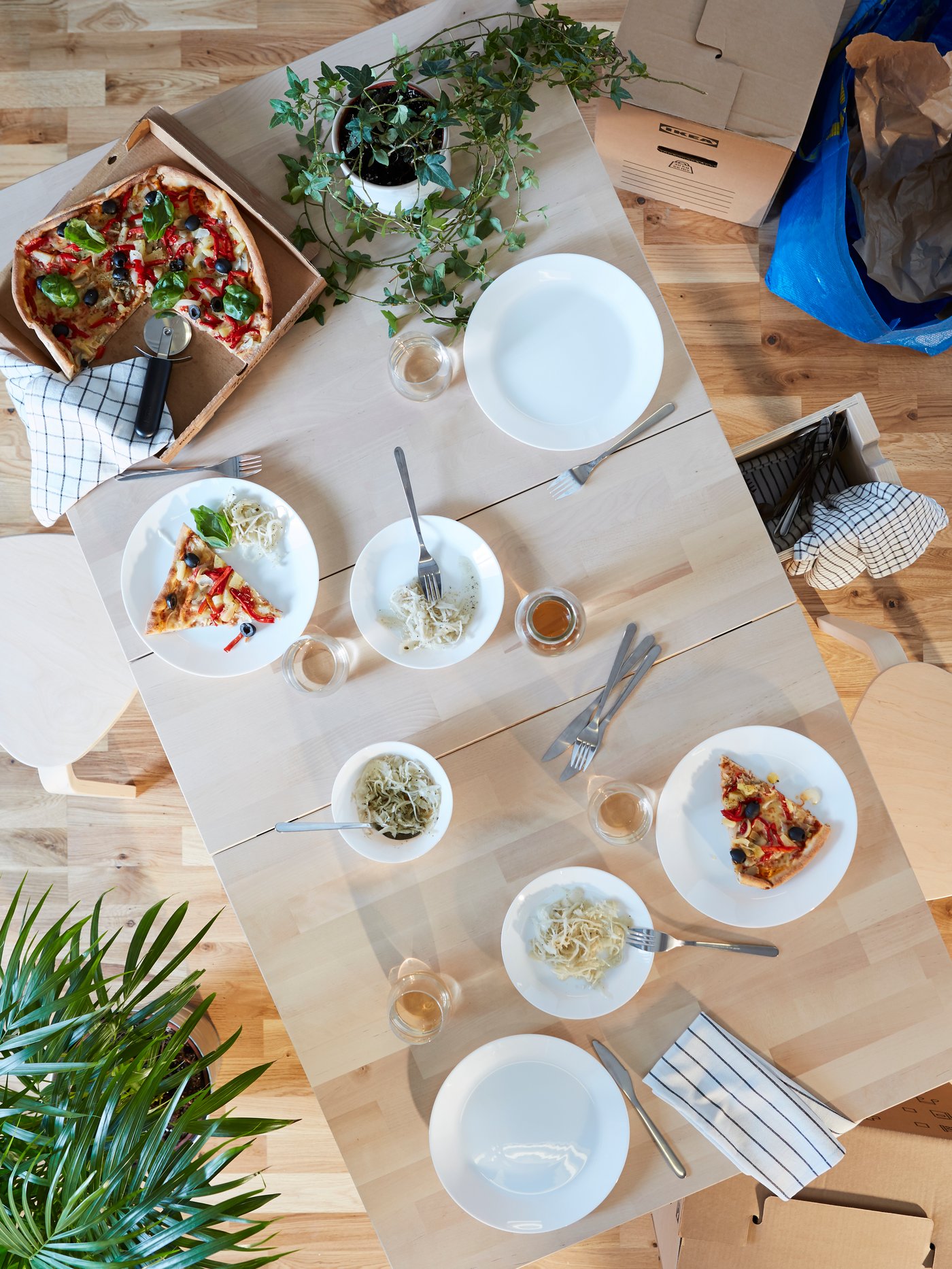 A bird's eye view of a birch dining table with IKEA OFTAST white dinnerware, a plant in a white pot and a pizza in a box.