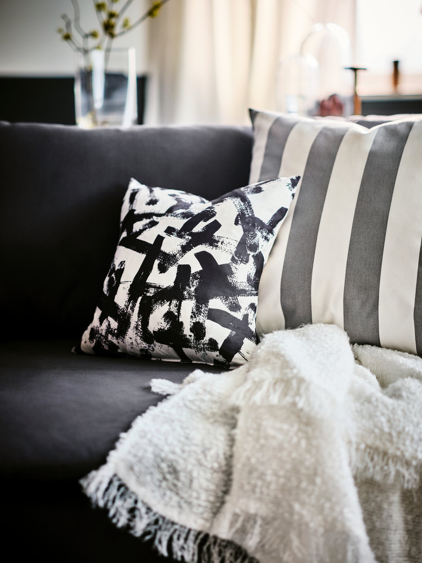 Un par de cojines IKEA sobre un sofá gris. Detrás se ven unas cortinas de color crema y una mesa con un florero encima.