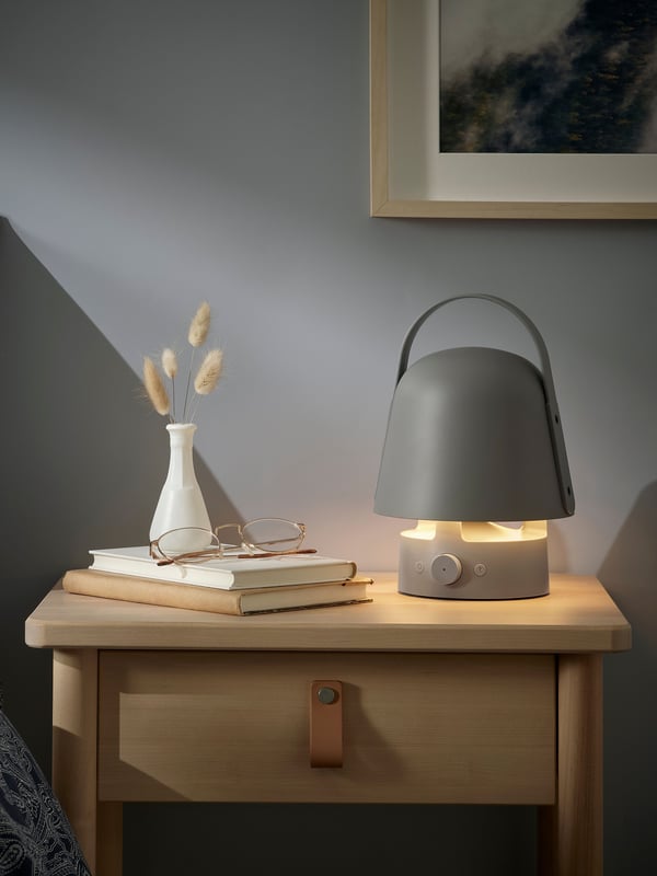 Siva VAPPEBY Bluetooth baštenska zvučnik-lampa u obliku pečurke s ručkom upaljena je i nalazi se pored knjiga na noćnom stočiću.