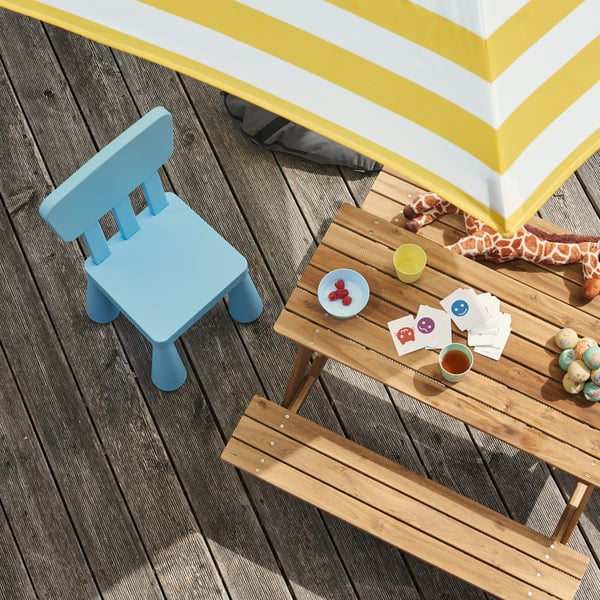 In a garden, a light blue MAMMUT children’s chair next to a wooden table with snacks, placed under a yellow parasol.
