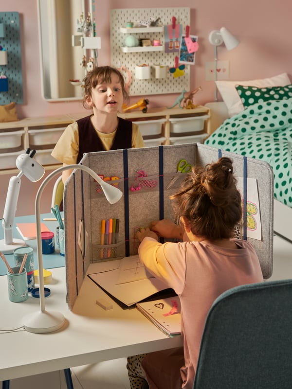 Two school girls sitting at their own desk facing each other with a desktop divider between them in a children’s bedroom.