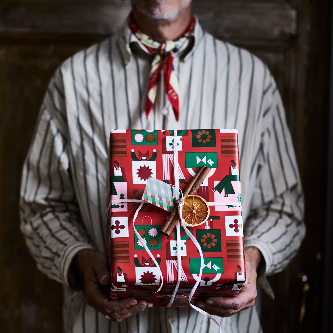 A person with a striped shirt is holding a present wrapped in VINTERFINT gift wrapper and adorned with a dried fruit.
