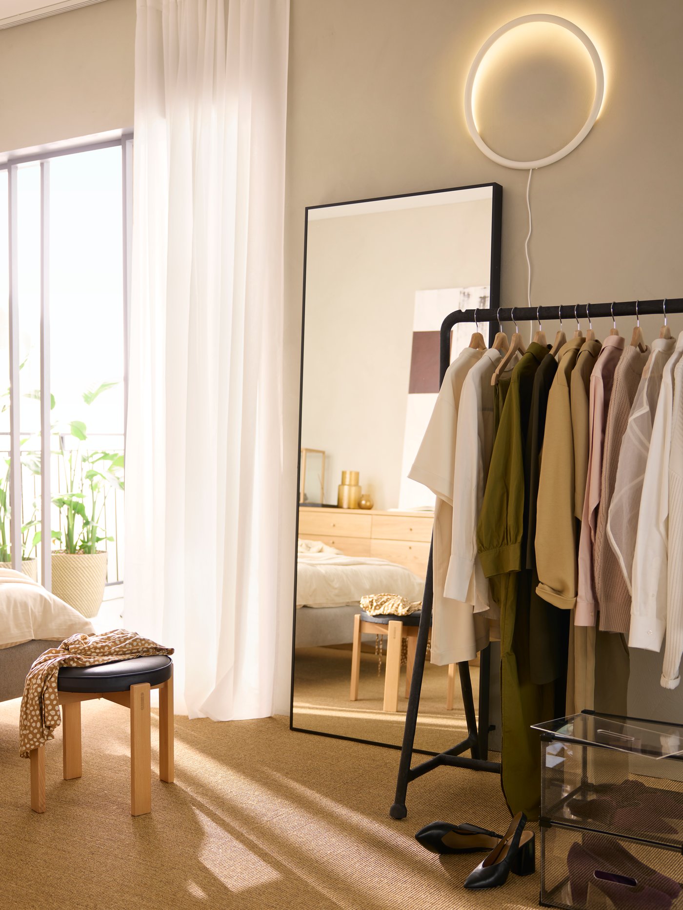A black TURBO clothes rack holding various items of clothing in a sunny room by a black HOVET mirror and a HOLMSJÖ stool.