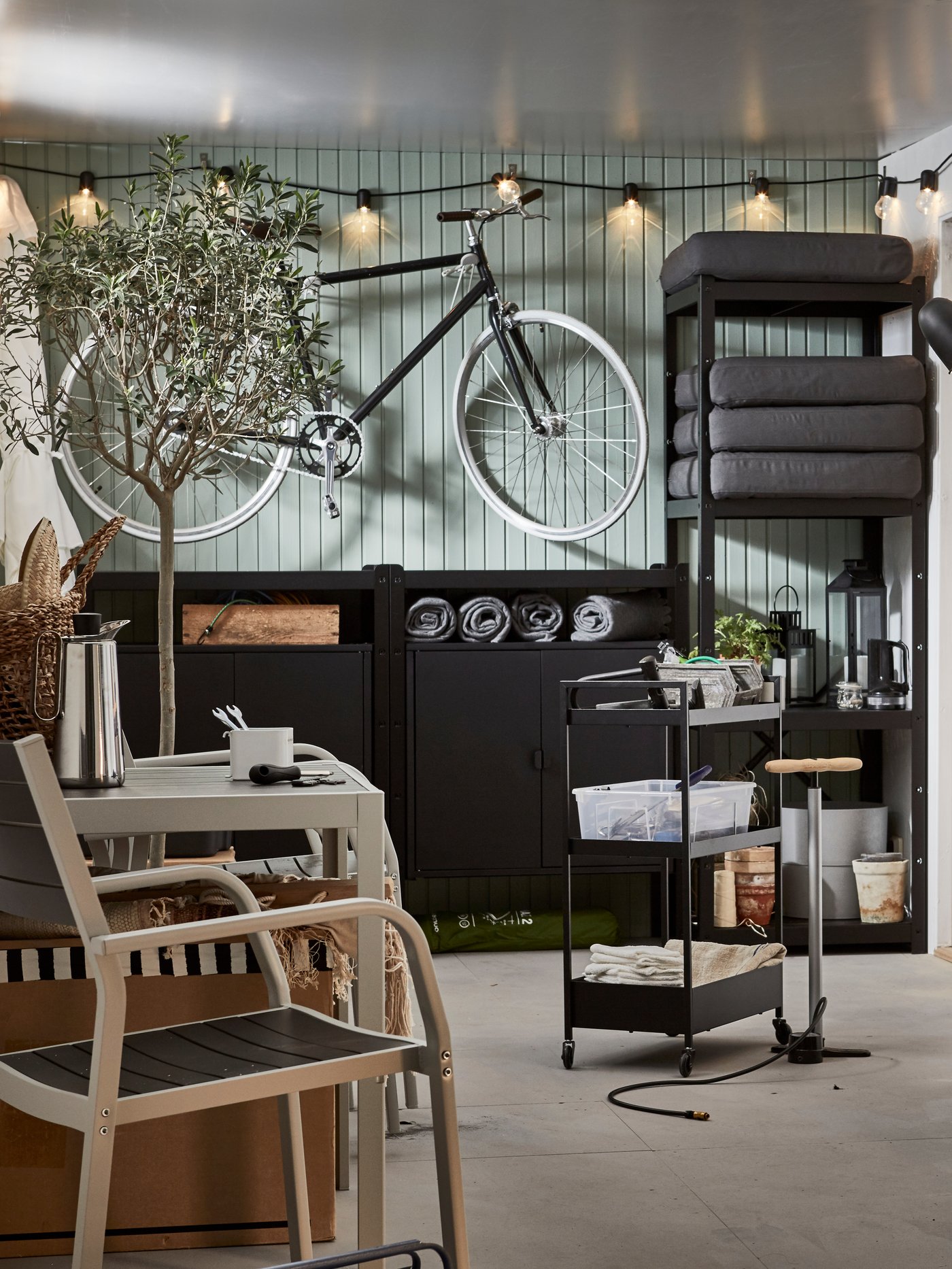A garage with bike, a lit lighting chain, a wall of storage, a seating area and a black NISSAFORS trolley holding tools.