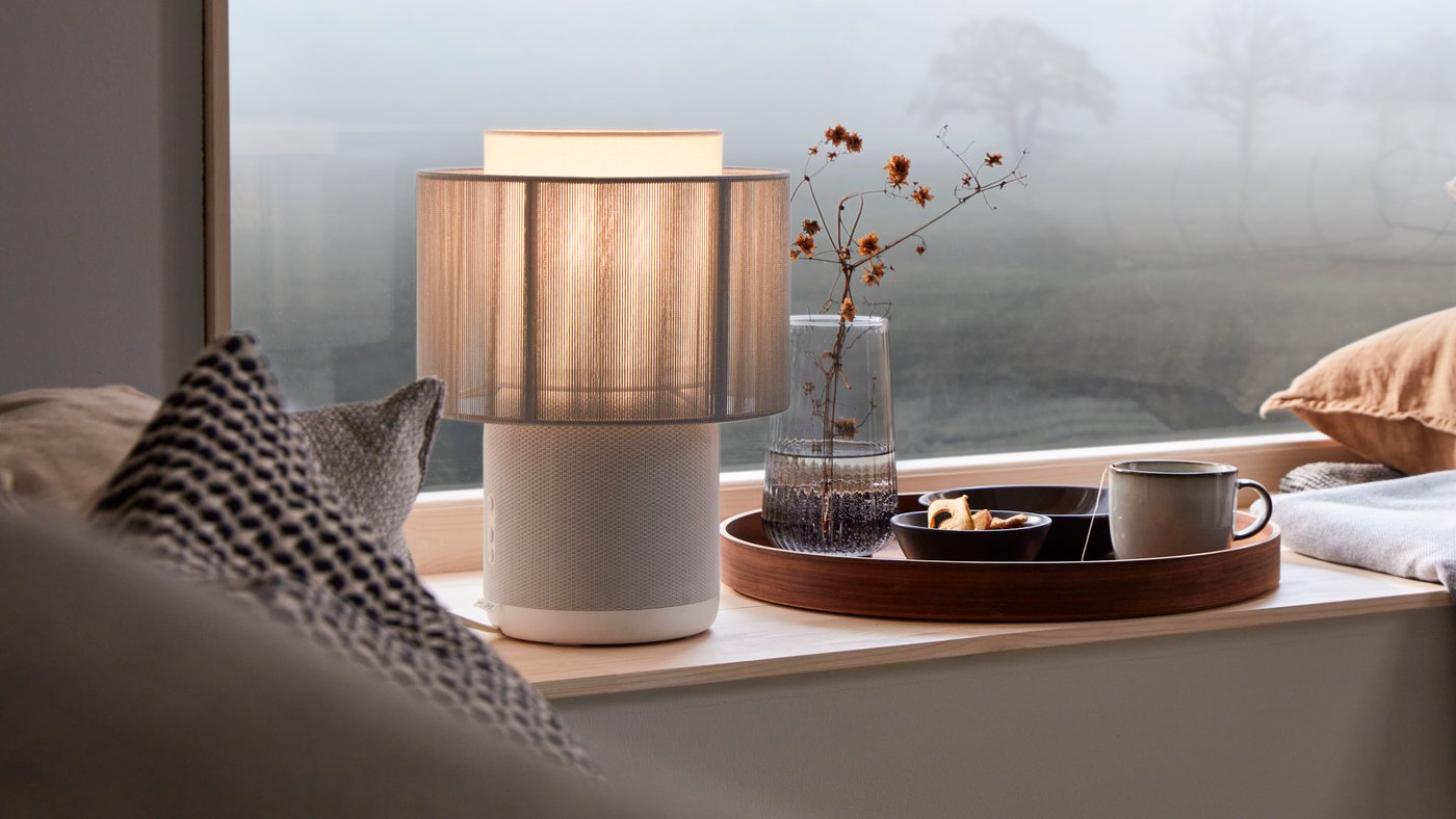 A lamp on the side of a window and a tray containing food, a flower, and a cup. Pillows can be seen in the bottom left-hand corner of the picture in the foreground.