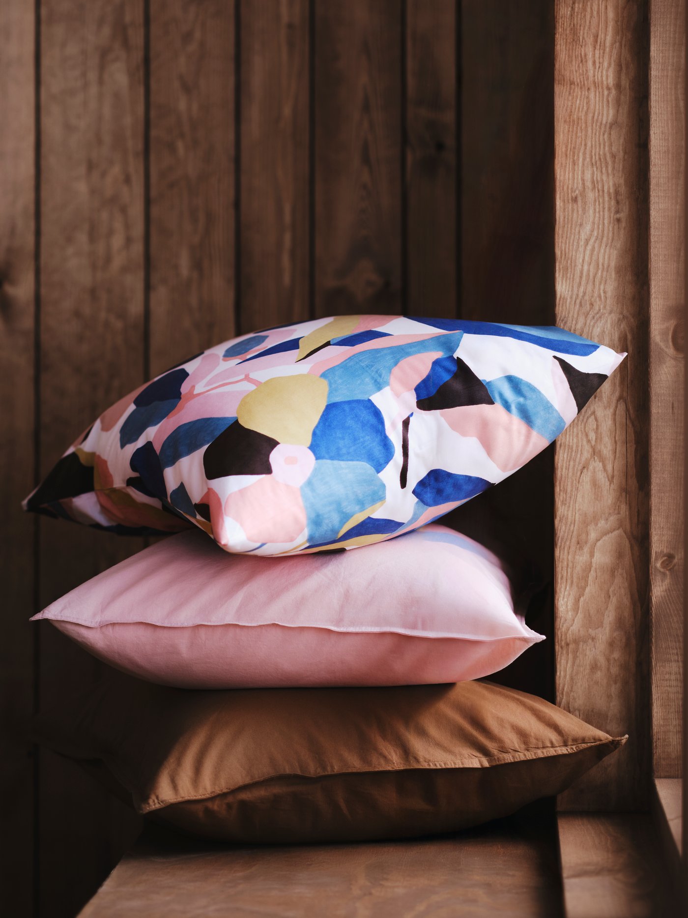 A stack of three pillows, one in a ÄNGSLILJA pillowcase in light pink, sit on a wooden bench against a wood-paneled wall.