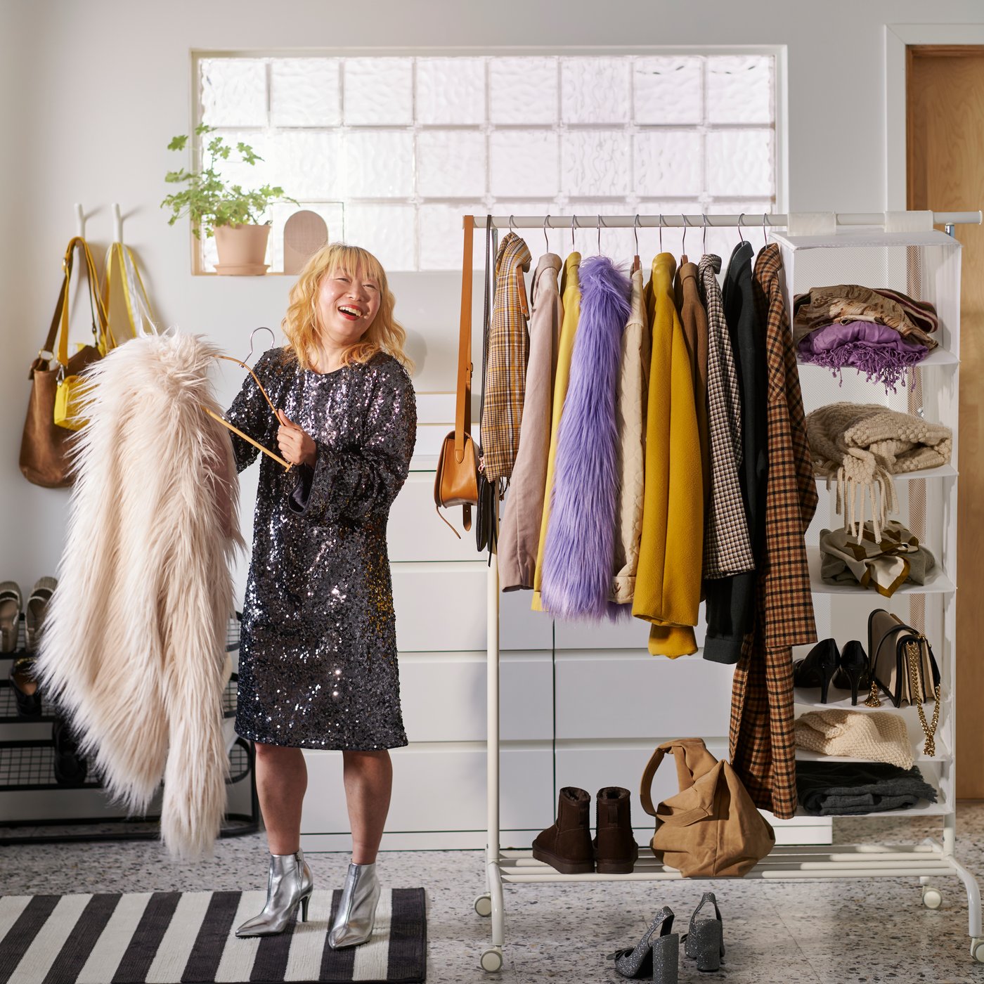 A woman in a hallway putting a coat on a hanger and standing beside a white RIGGA clothes rack with coats and shoes on.