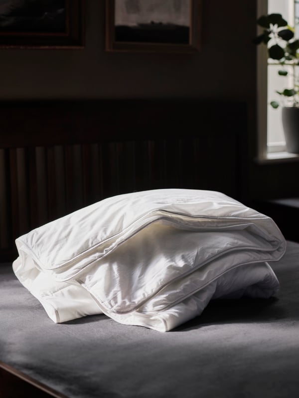 A STJÄRNBRÄCKA warm duvet folded up on a grey mattress in a dark room near a window with a plant on the sill.