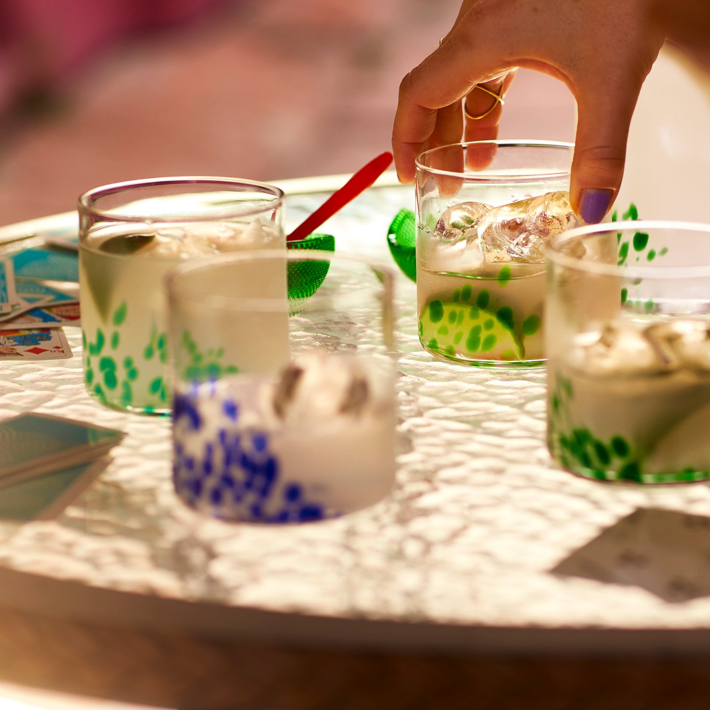 Four ÖMSESIDIG glasses in clear glass with mixed colours on a small glass table. A hand is putting one of the glasses down.