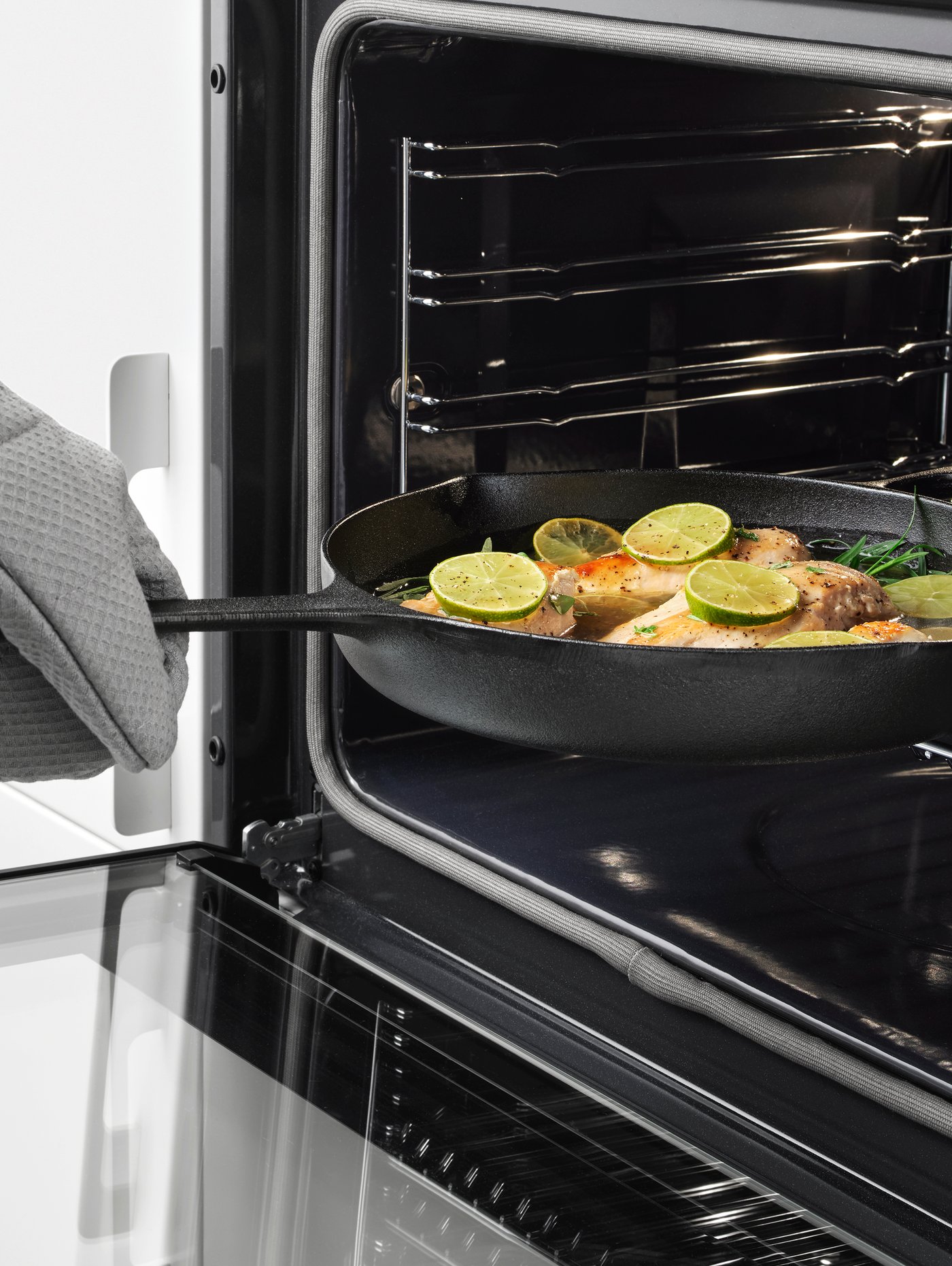 A hand wearing an oven glove placing a VARDAGEN cast iron frying pan containing fish, lime, and herbs into an oven.
