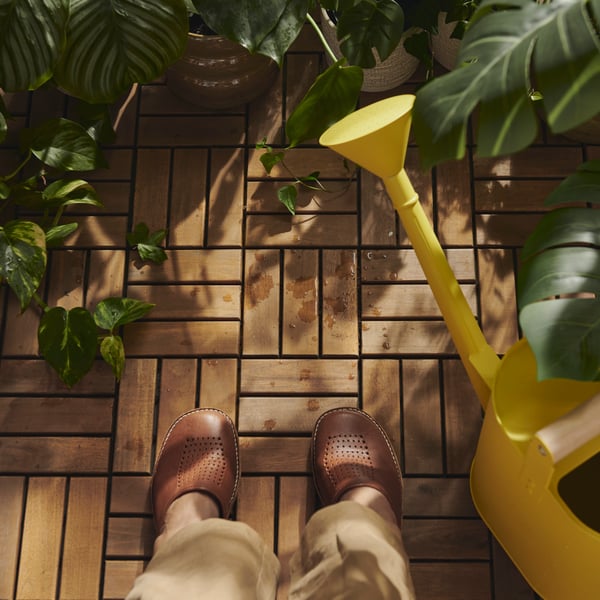 A balcony with acacia RUNNEN outdoor floor decking and a pair of shoes, surrounded by green plants and a yellow watering can.
