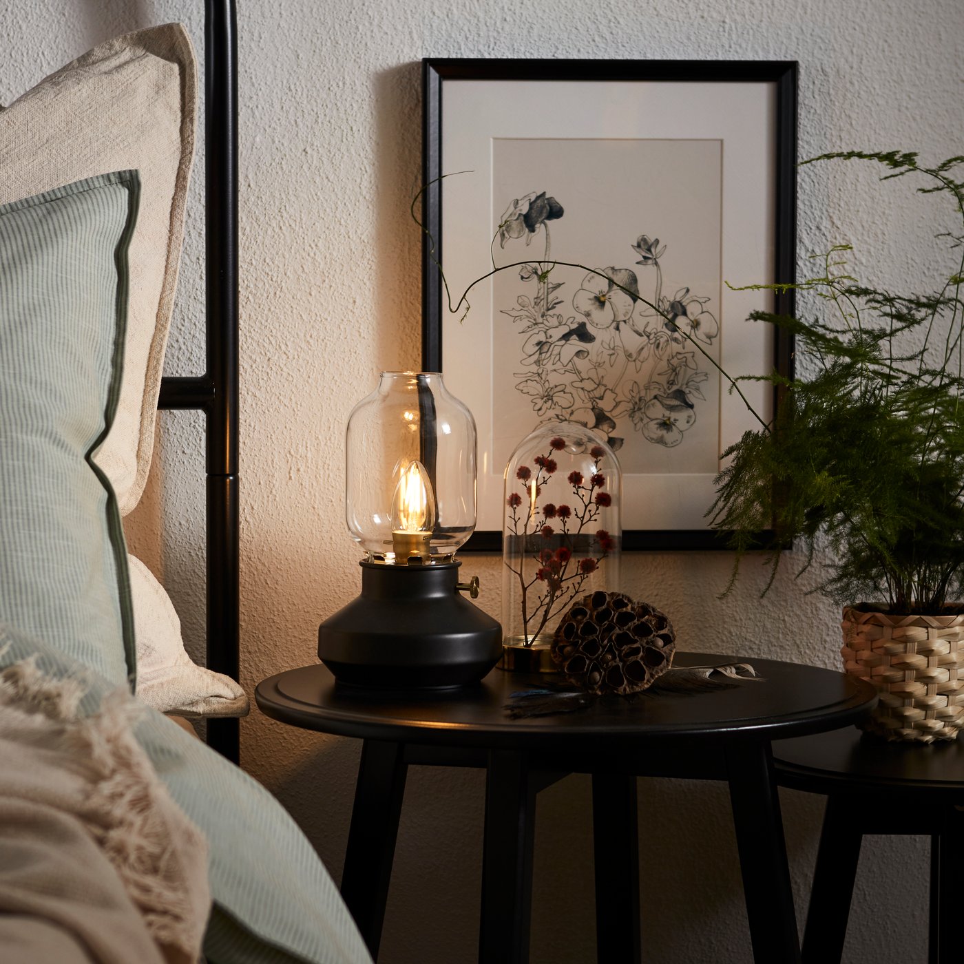 Edge of made bed with high headboard, round bedside table with an LED lamp in the form of a kerosene lamp.