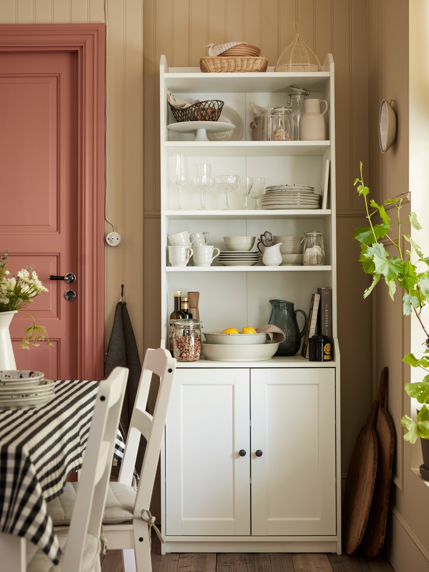A HAUGA high cabinet with 2 doors holding tableware and food stands in a corner of a dining area near a table and chairs.