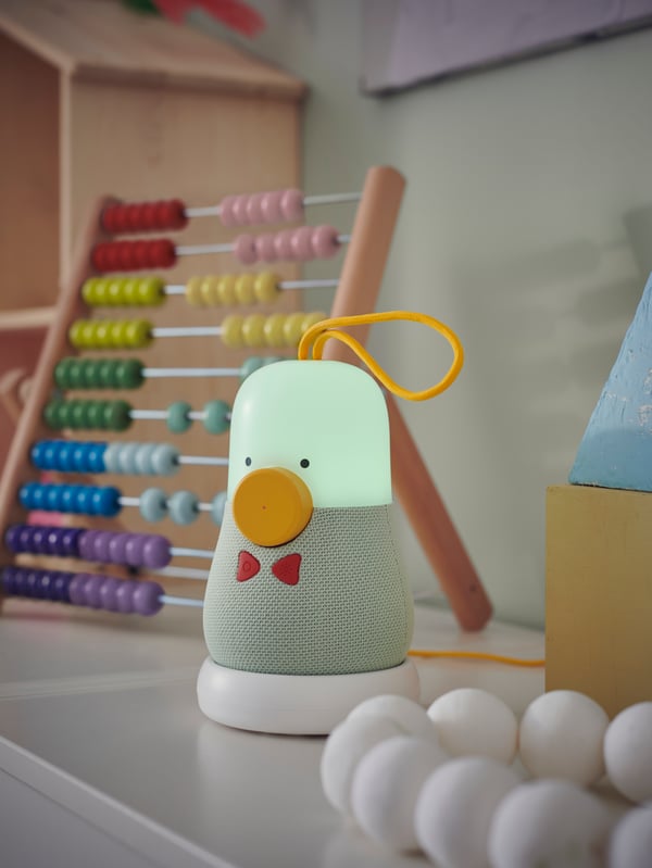 A green VAPPEBY peanut shape Bluetooth speaker with the light on stands near an UNDERHÅLLA abacus on a white surface.