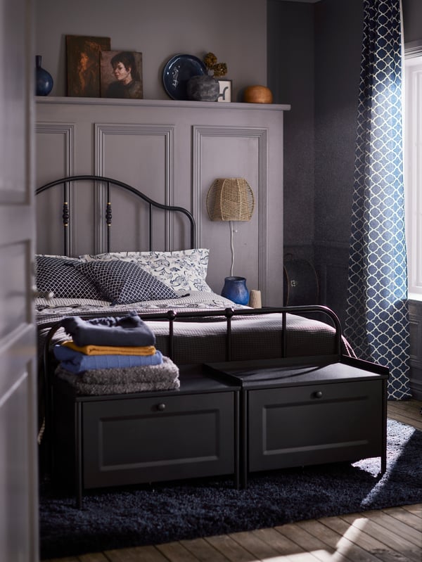 Two black storage benches and a bed with a black SAGSTUA bed frame made with white/blue floral bed linen on a dark blue rug.