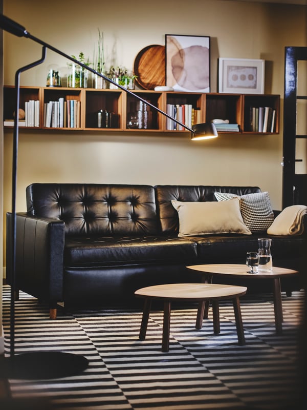 A black leather LANDSKRONA sofa sits on a black and white striped rug with two round coffee tables. Shelving is overhead.