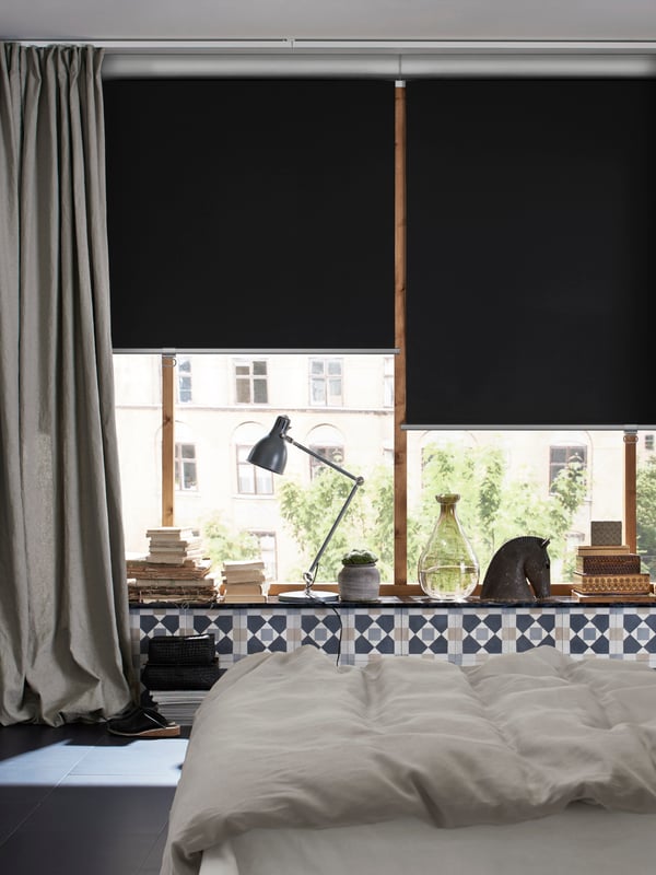 A bed with plenty of pillows, plants all around, a window with books, magazines, plants and a block-out roller blind.