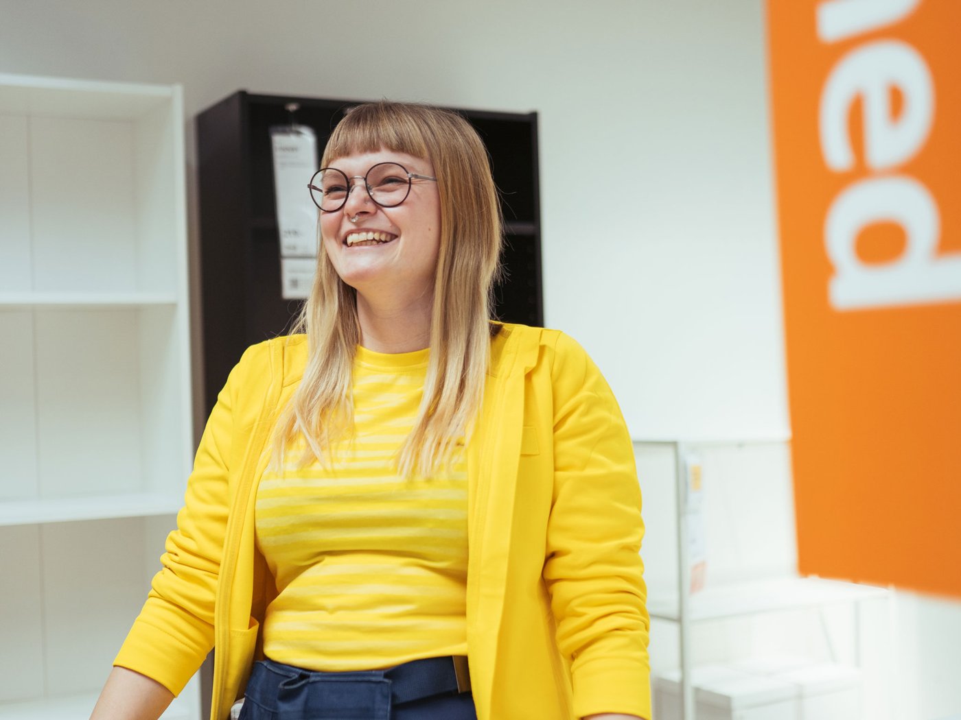 En person med langt blond hår og briller i en gul trøje står i et IKEA varhus og smile bredt