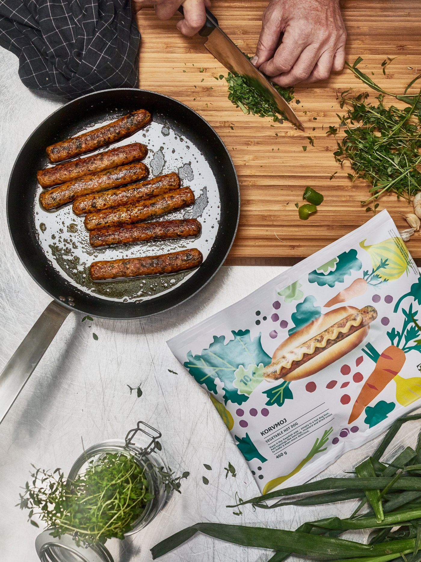 A person chops sprigs of thyme to be added to a freshly fried pan of veggie hot dogs, besides which is the KORVMOJ packet.