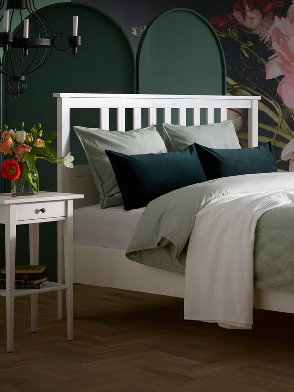 Katil HEMNES berwarna putih diletakkan di sebelah meja sisi katil HEMNES berwarna putih yang terdapat vas dengan bunga di atasnya.