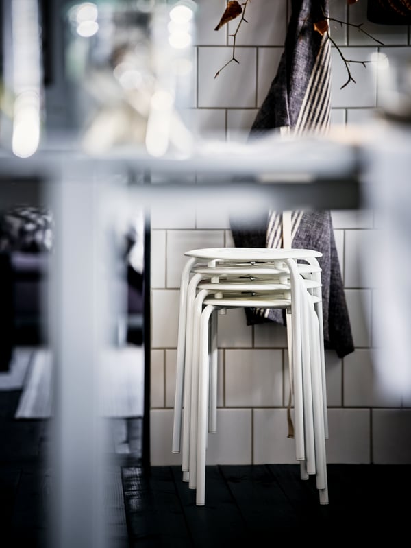 Stacked white MARIUS stools stand by a white-tiled wall in a black-and-white-themed kitchen.