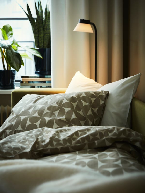 Partie d’un canapé-lit causeuse SKÖNABÄCK jaune-vert utilisé comme lit, avec du linge de lit ÄNGSNEJLIKA gris-beige et blanc cassé.
