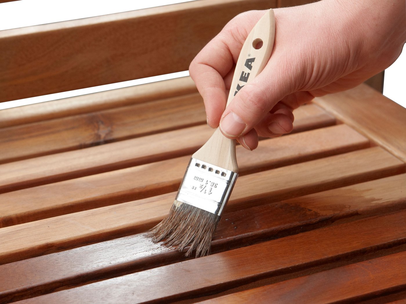 Primer plano de una mano con un pincel que barniza una superficie de madera. Las cerdas del pincel están en contacto con la madera y aplican el líquido de manera uniforme. La superficie de madera se compone de varias tablas, lo que parece indicar que se trata de un mueble o de un piso de madera.