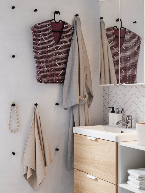 A group of black hooks arranged in a graphic way on the bathroom wall, with a towel, bathrobe, blouse and necklace.