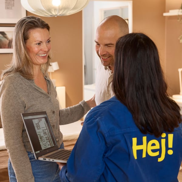 Standing in a showroom with light brown walls, an IKEA interior designer is showing two of her clients interior drawings on a laptop.